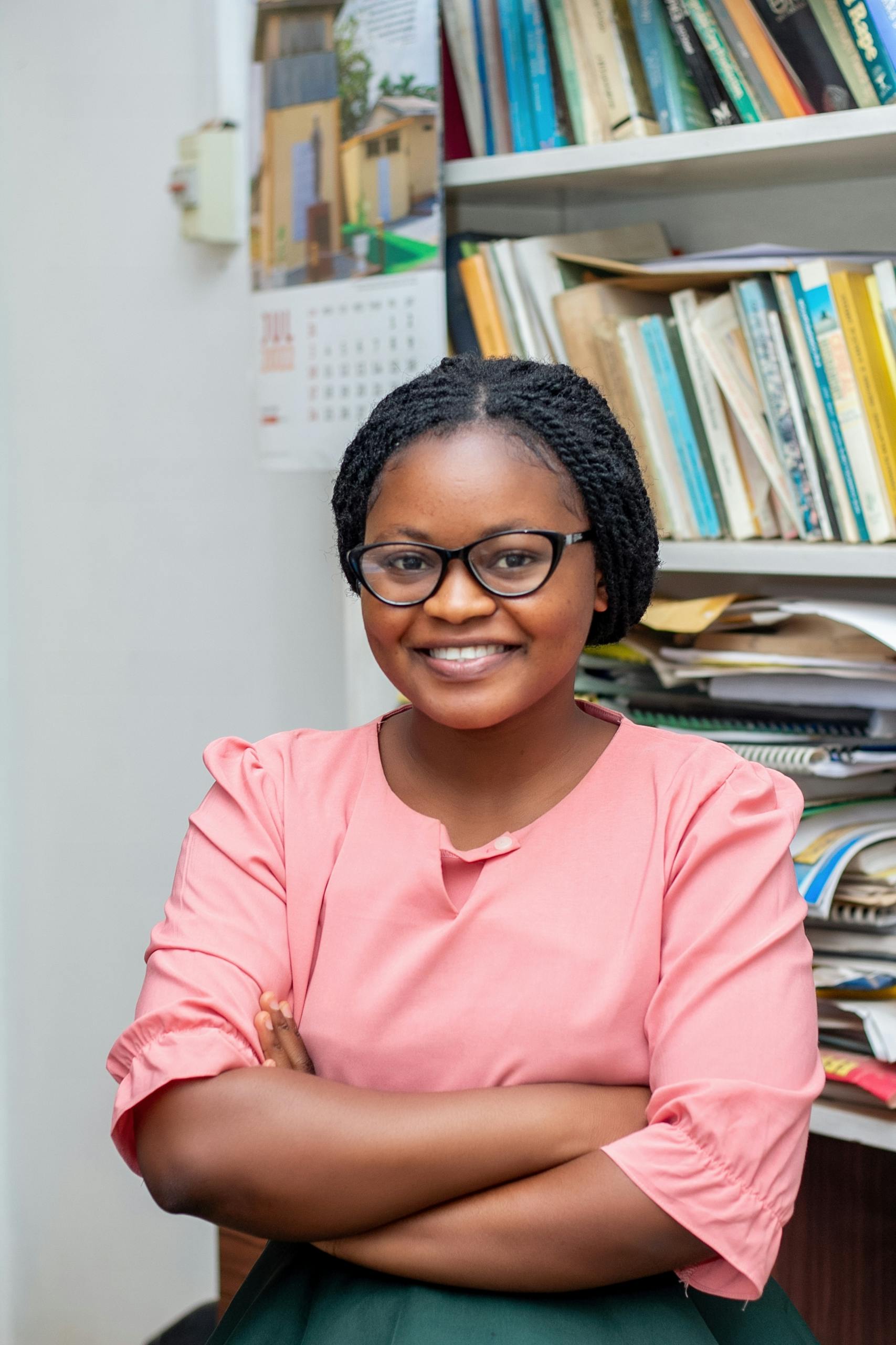 Portrait of a black woman smiling confidently with crossed arms in an educational setting with book shelves.